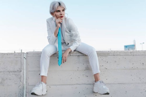 thoughtful young woman wearing white suit sitting on retaining wall against clear sky - fashion stock-fotos und bilder