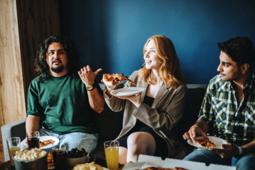 three cheerful gen z friends sitting on sofa in cozy room eating pizza and talking - junk food stock pictures, royalty-free photos & images