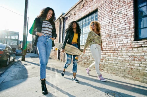 three young women with long curly hair walking along pavement, one carrying skateboard. - fashion stock pictures, royalty-free photos & images