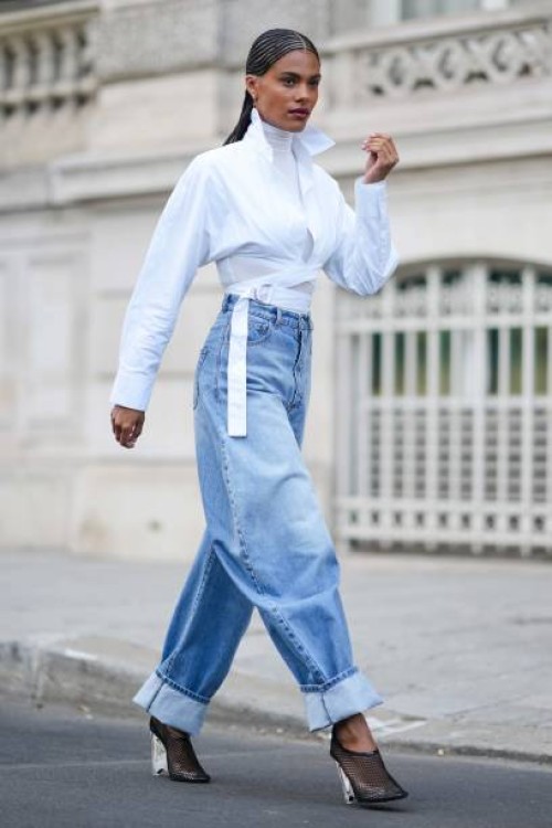 Tina Kunakey wears gold earrings, a white turtleneck long sleeves t-shirt, a white laces waist cropped waist shirt, high waist blue faded denim puffy...