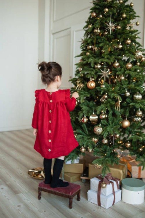 toddler girl in red linen dress and a headband decorates christmas tree at home. christmas outfit. preparing for celebration. festive season. lifestyle portrait. - home decoration stock pictures, royalty-free photos & images