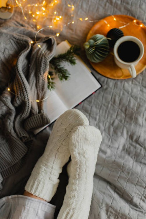 top view of female feet in cozy white socks on bed with cup of coffee, christmas lights and open book. christmas, relaxation and hygge concept. - home decoration stock pictures, royalty-free photos & images