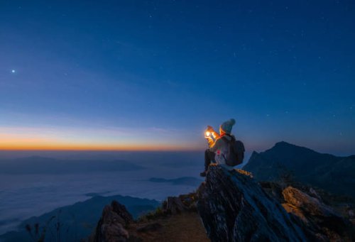 tourist woman holding a lantern while sitting on the rock on top of doi pha tang mountain a high cliff over the thai-laotian border located in wiang kaen district of chiang rai province of thailand. beautiful view before the 