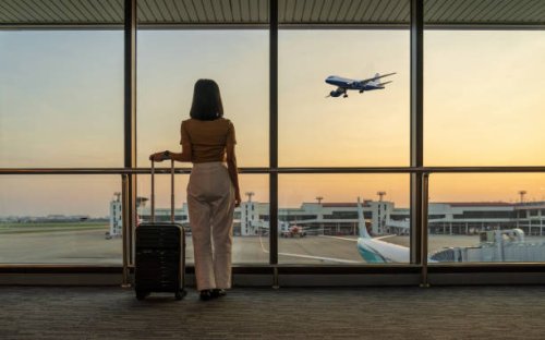 travel tourist standing with luggage watching sunset at airport window. woman looking at lounge looking at airplanes while waiting at boarding gate before departure. travel lifestyle. transport and travel concept - travel sto