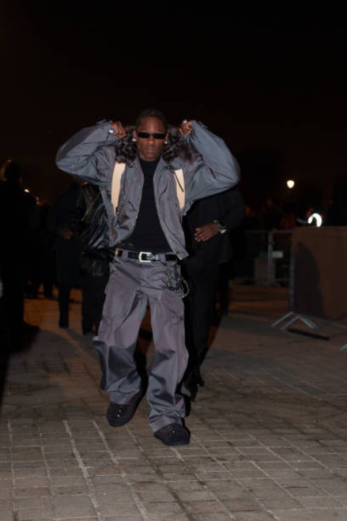 Travis Scott wears sunglasses, a gray bomber jacket with fluffy faux fur collar, a black t-shirt, a leather belt, cargo pants, before the Louis...