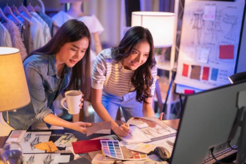two asian female cloth designer discussion at night brainstorm new ideas,asian partners team looking at new deign sketch collection on table at home studio,.working late creative digital development agency - fashion stock pic