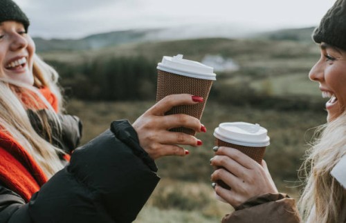 two beautiful young woman hold cardboard coffee cups and make a celebratory toast by bumping them together - junk food stock pictures, royalty-free photos & images