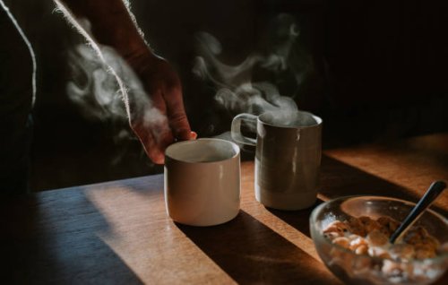 two cups of coffee on a wooden table with a bowl of breakfast cereal. someone lifts one of the mugs out of frame. - coffee stock pictures, royalty-free photos & images