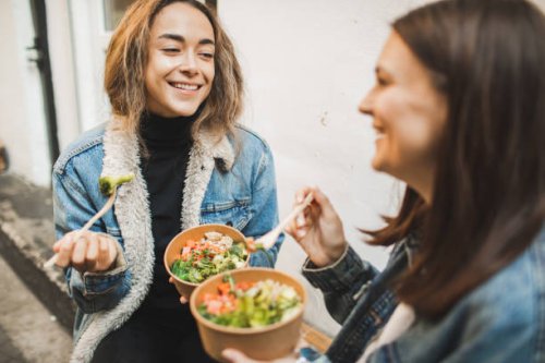 two females friends eating take away vegetarian bowls outdoors in town. - junk food stock pictures, royalty-free photos & images
