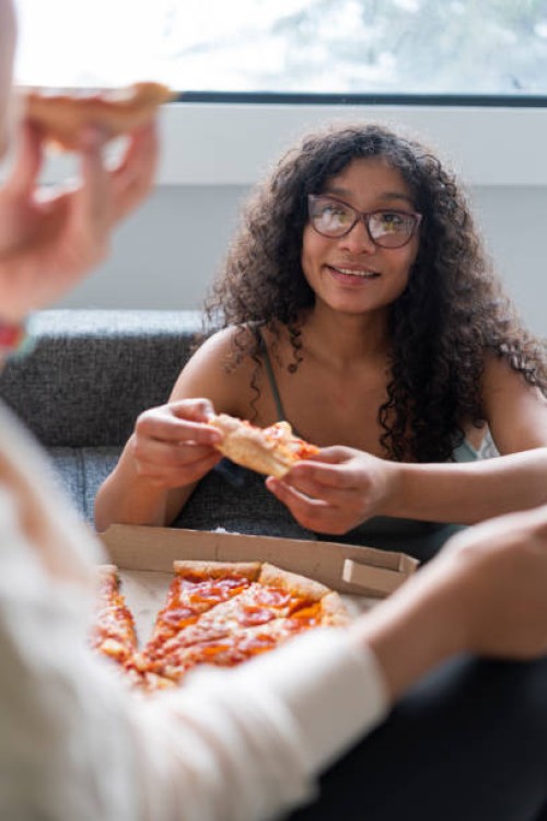 two latin friends spend time together in the living room of their house eating a piece of pizza - junk food stock pictures, royalty-free photos & images