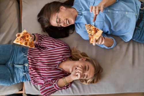 two laughing young women lying down eating pizza together - food stock pictures, royalty-free photos & images