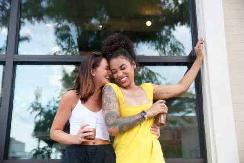 two millennial women, one caucasian with shoulder length hair and the other mixed race with curly hair, laugh while they hug each other outdoor near a building, wearing casual summer clothing. - fashion stock pictures, royalt