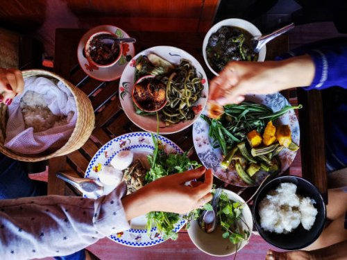 two thai women traditionally eating a selection of freshly cooked northern thai food of vegetables, soups and curries served in dishes on a wooden table, along with sticky rice, which is a food staple in northern and northeas