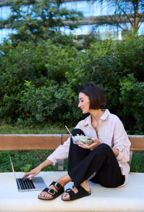 vertical photo of a businesswoman sitting on an outdoor bench, eating a healthy salad during her lunch break while working on a laptop computer - junk food stock pictures, royalty-free photos & images