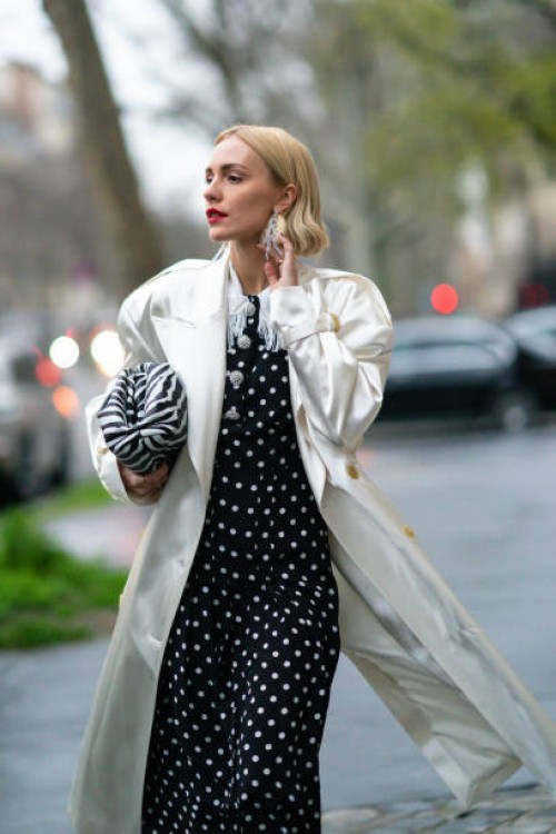 Viktoria Rader wears long bejeweled earrings, a white long coat, a black and white zebra print puff bag, a black and white dress with polka dots,...