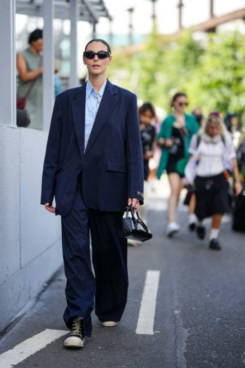 Vittoria Ceretti wears sunglasses, a blue shirt, a navy blue oversized blazer jacket, wide leg flared suit pants, sneakers, a black leather bag,...
