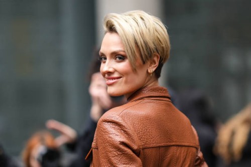 Wallis Day wears gold earrings, a camel shiny leather long coat , outside David Koma, during London Fashion Week February 2023 on February 18, 2023...