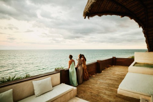 wide shot of laughing female friends standing on deck of luxury hotel suite looking at ocean and sunset - fashion stock pictures, royalty-free photos & images