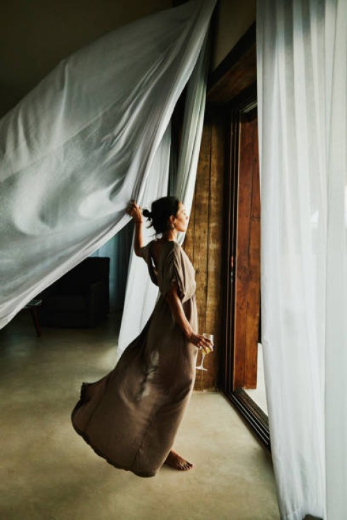 wide shot of woman standing by door of luxury hotel suite looking out at view with curtains blowing in wind - fashion stock pictures, royalty-free photos & images