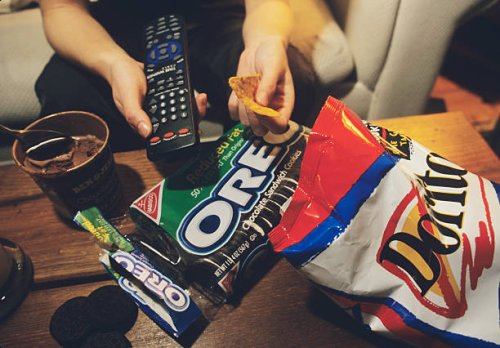 With remote control in one hand and nacho chip in the other, a teenager eats the food she likes in front of the TV.