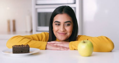 woman choosing between a cake or apple in the kitchen to eat as a day time snack at home. indian girl thinking about the healthy, nutrition and organic fruit or the unhealthy chocolate dessert. - junk food stock pictures, roy