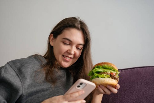 woman eating a big burger with meat, tomatoes and lettuce sitting on the couch at home, looking at the phone and smiling, concept of fast food - junk food stock pictures, royalty-free photos & images