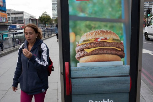 Woman eating a burger while passing an advertisement for burgers at Elephant and Castle in London, UK. The area is now subject to a master-planned...
