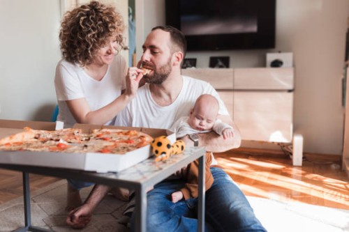 woman feeding pizza to her husband who is holding a sleeping baby - junk food stock pictures, royalty-free photos & images