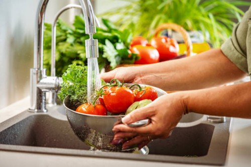woman holding a colander full of fresh vegetables under running water in kitchen sink - food stock pictures, royalty-free photos & images