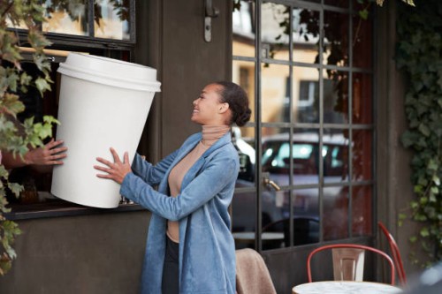 woman holding large disposable coffee cup at take away counter of cafe - junk food stock pictures, royalty-free photos & images