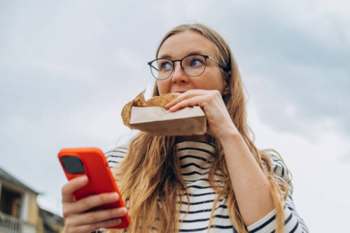 woman holds a smartphone in her hands and eats a bun with appetite on a city street - junk food stock pictures, royalty-free photos & images