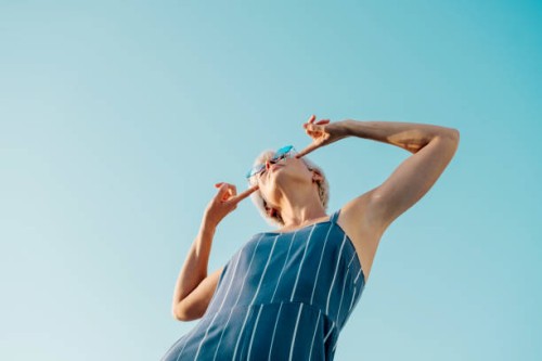 woman in a blue jumpsuit striking a playful pose, pointing at her stylish glasses, under a bright blue sky, radiating fun and confidence. - fashion stock pictures, royalty-free photos & images