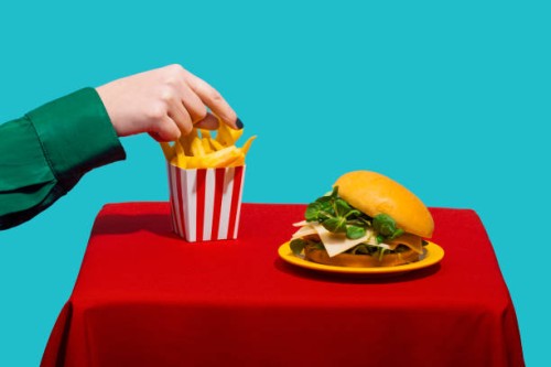 woman reaching for french fries near cheeseburger on table against blue background - junk food stock pictures, royalty-free photos & images