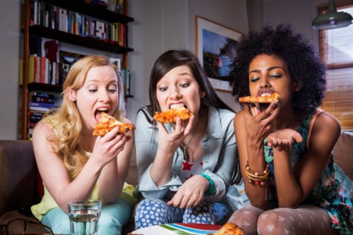 women biting into pizza sitting together on sofa. - junk food stock pictures, royalty-free photos & images