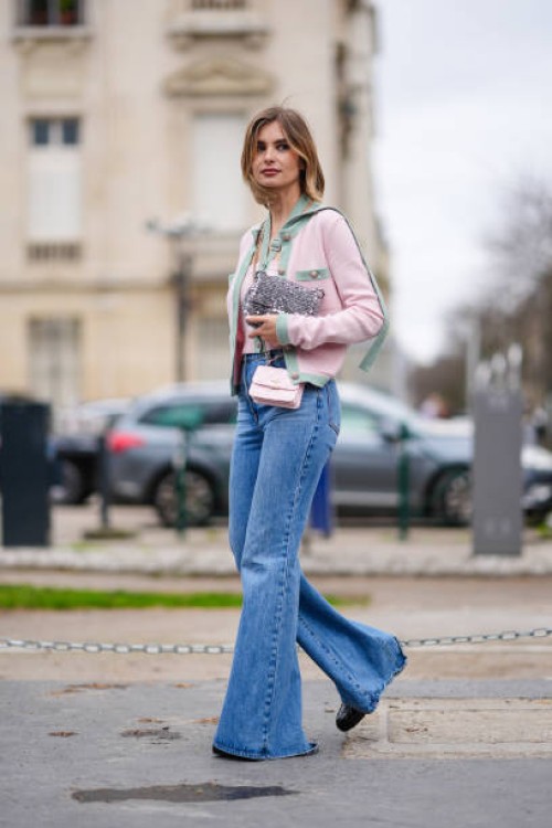 Xenia Adonts wears a pastel pale green and pink jacket , a silver glitter shiny bag, a pink mini bag, blue flared denim pants / jeans, outside...