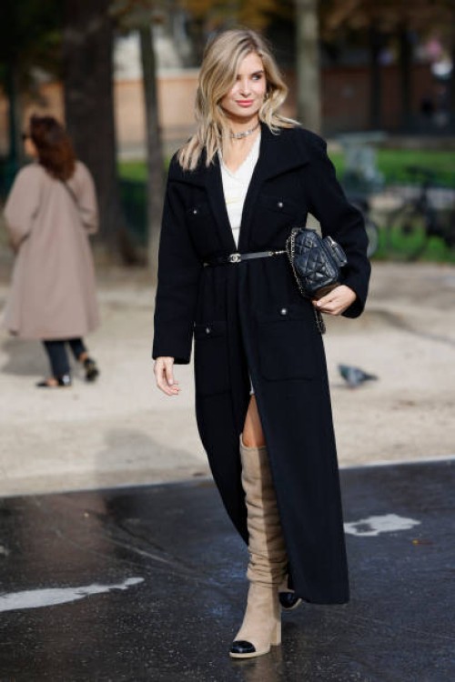 Xenia Adonts wears white mini dress, black long coat, black Chanel belt, black Chanel bag, beige over the knees Chanel boots, outside Chanel, during...