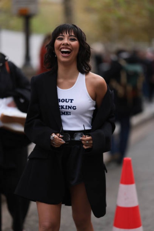 Xochitl Gomez is seen wearing an oversized black blazer with peak lapels and shoulder pads, a white tank top with a dark blue printed quote saying...