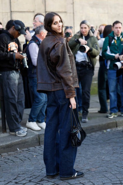 Yasmin Wijnaldum wears brown leather jacket, blue wide leg jeans, Adidas black sneakers, black bag, outside Nina Ricci, during the Womenswear...
