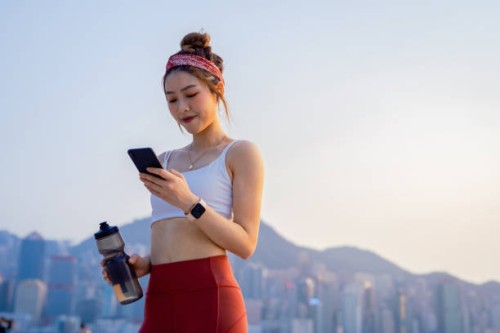 young asian sports woman using smartphone after exercising outdoors in city in the morning, against urban city skyline. relaxing after running. youth culture. habits and hobbies. active lifestyle. health and fitness with tech