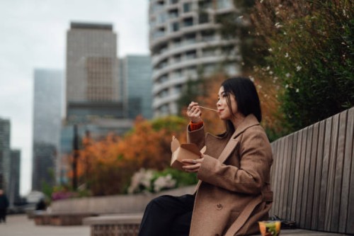 young asian woman eating takeaway lunch while sitting on the bench in city park - junk food stock pictures, royalty-free photos & images