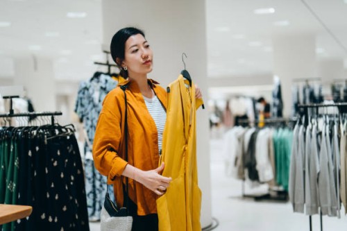 young asian woman shopping in a clothing store, looking at a yellow dress in mirror. sale season. seasonal shopping - fashion stock pictures, royalty-free photos & images