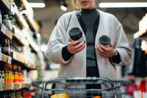 young asian woman with shopping cart shopping for cooking sauce in supermarket, reading and comparing the product information of two bottles of cooking sauces. routine grocery shopping. making healthier food choices. consumer