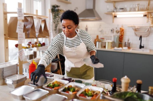 young beautiful female chef packing food in aluminum plates, getting it ready for delivery at her organic food store - food stock pictures, royalty-free photos & images