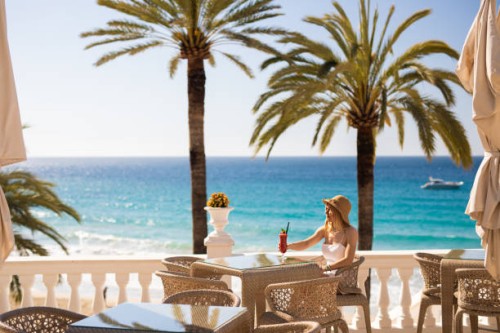 young blonde woman sitting in the beach restaurant enjoying a red cocktail with the turquoise sea and palm trees and a yacht in the background - travel stock pictures, royalty-free photos & images
