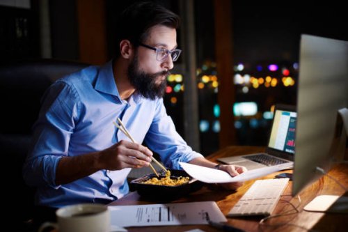 young businessman looking at computer and eating takeaway at office desk at night - junk food stock pictures, royalty-free photos & images