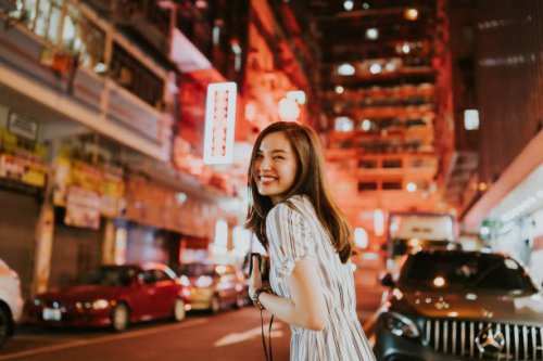 young cheerful asian woman looking back in front of the neon lights of hong kong on the streets at night while holding her camera - travel stock pictures, royalty-free photos & images