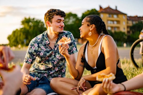 young couple look at each other in love while sitting in a park eating pizza - food stock pictures, royalty-free photos & images