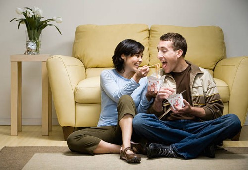 young couple sitting cross-legged in front of sofa eating takeaway food, woman feeding man with chopsticks, at home - junk food stock pictures, royalty-free photos & images