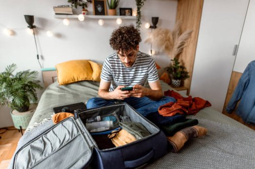 young man sitting on his bed, using smart phone and packing suitcase - travel stock pictures, royalty-free photos & images