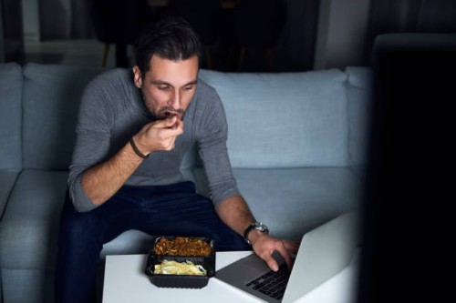 young man sitting on sofa in evening eating takeaway and using laptop - junk food stock pictures, royalty-free photos & images